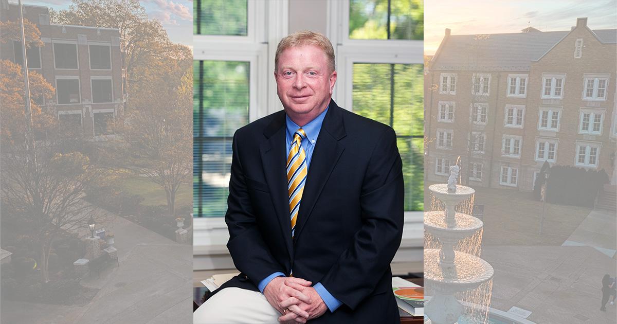 Portrait photo of College of Business and Technology Dean Greg Carnes sitting on the edge of his desk with his hands folded.